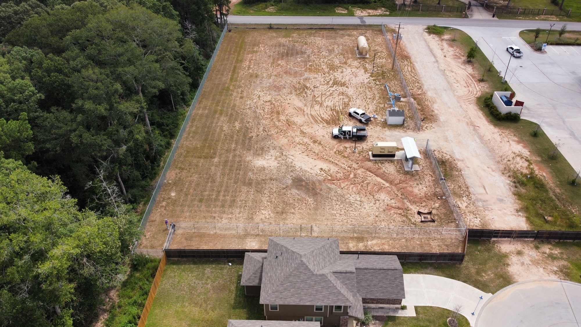 Aerial view of a fenced utility site beside a wooded area before major construction begins